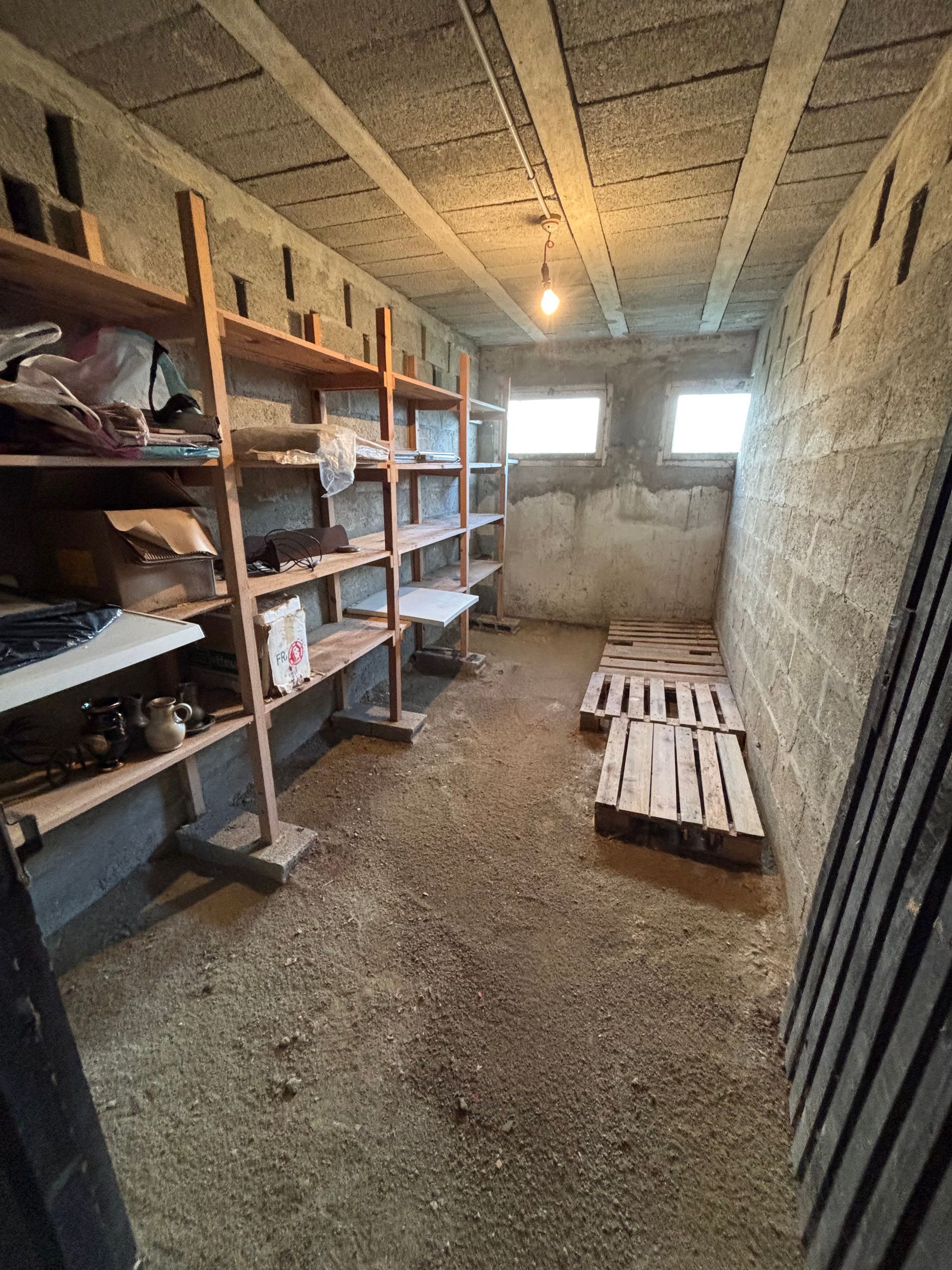 Un débarras aux murs en parpaings, avec des étagères en bois, un sol nu et une simple ampoule suspendue.