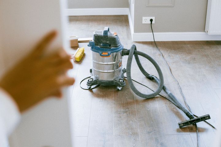 Aspirateur industriel sur un sol en bois près d'un mur avec une prise électrique ; une main est au premier plan.