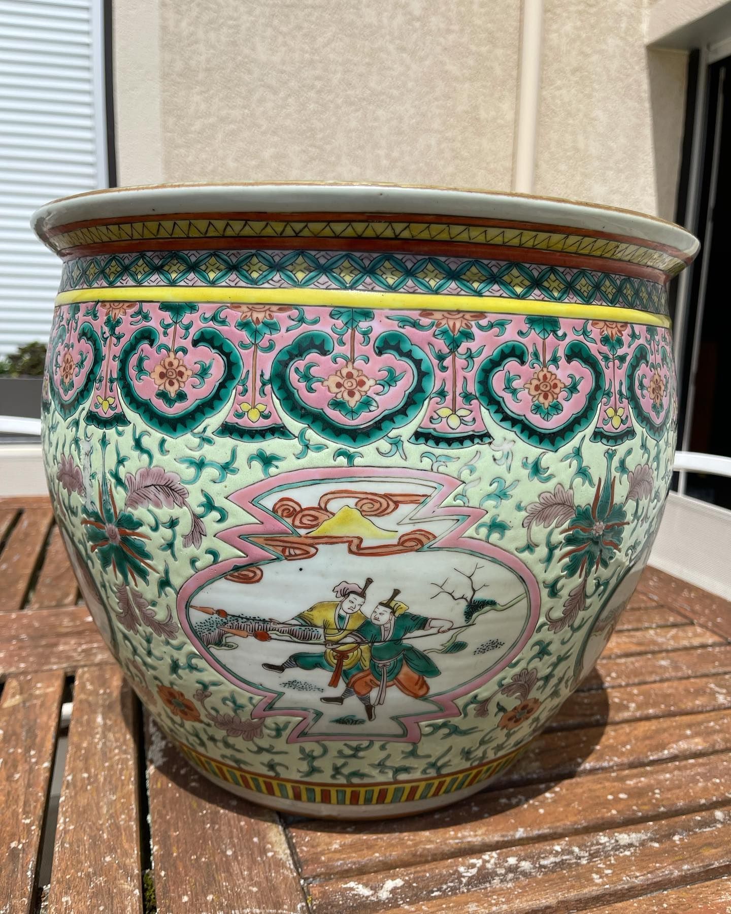 Grand pot de fleurs en porcelaine chinoise richement orné de motifs floraux et figuratifs roses, verts et jaunes, posé sur une table en bois.