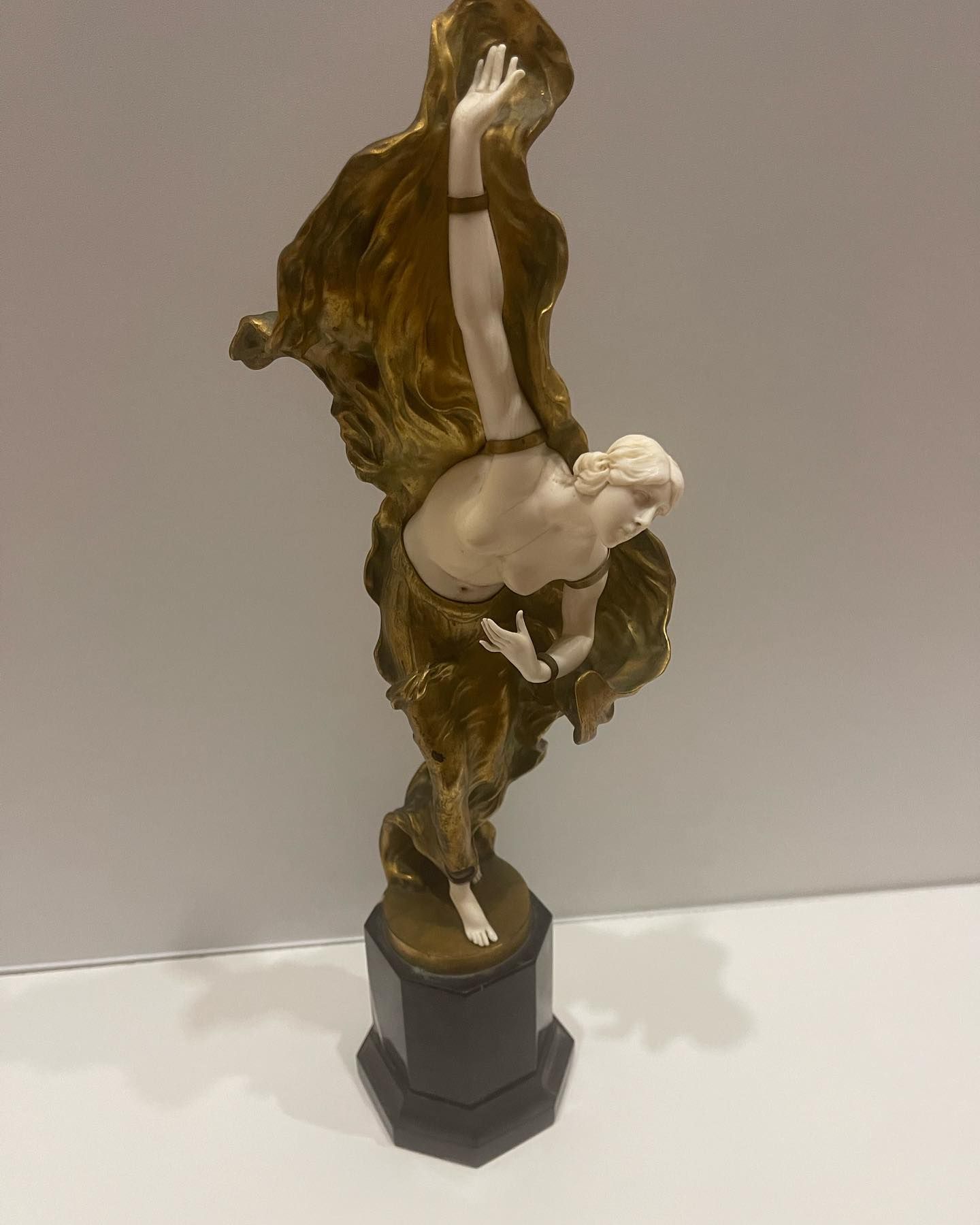 Statue d'une figure nue drapée d'un tissu doré, posant sur un socle hexagonal sombre, tendant les bras vers le ciel.
