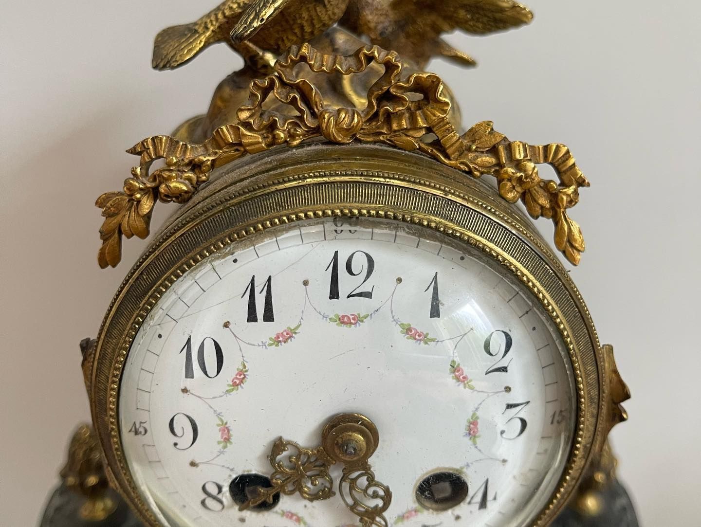 Gros plan sur le cadran décoratif d'une horloge. Cadran blanc à chiffres noirs, motifs floraux, boîtier doré et oiseau sur le dessus.