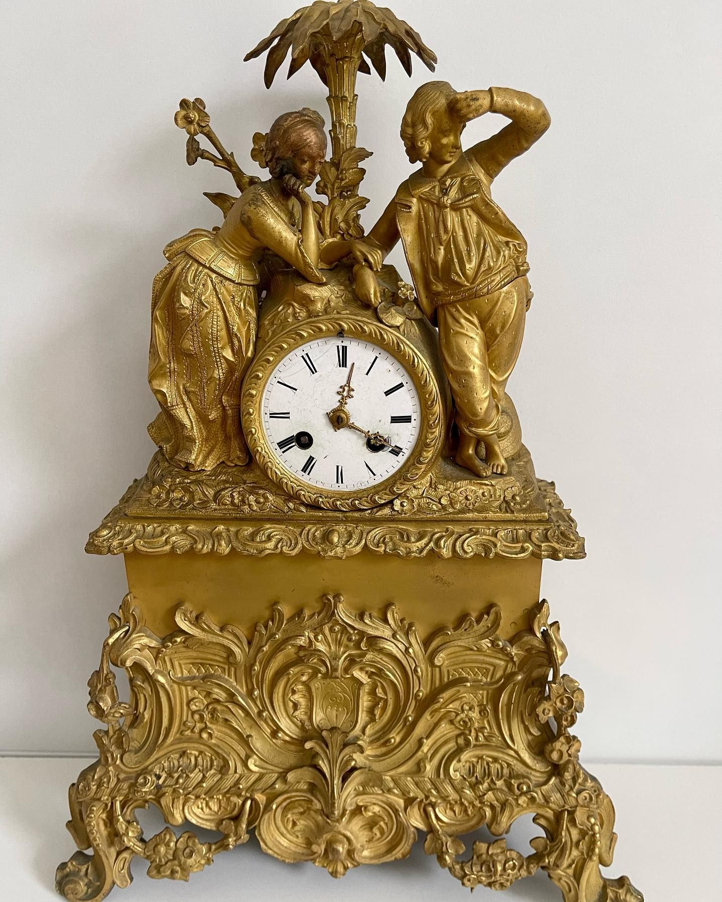 Horloge ancienne de couleur dorée avec deux personnages, des détails complexes et un cadran circulaire.