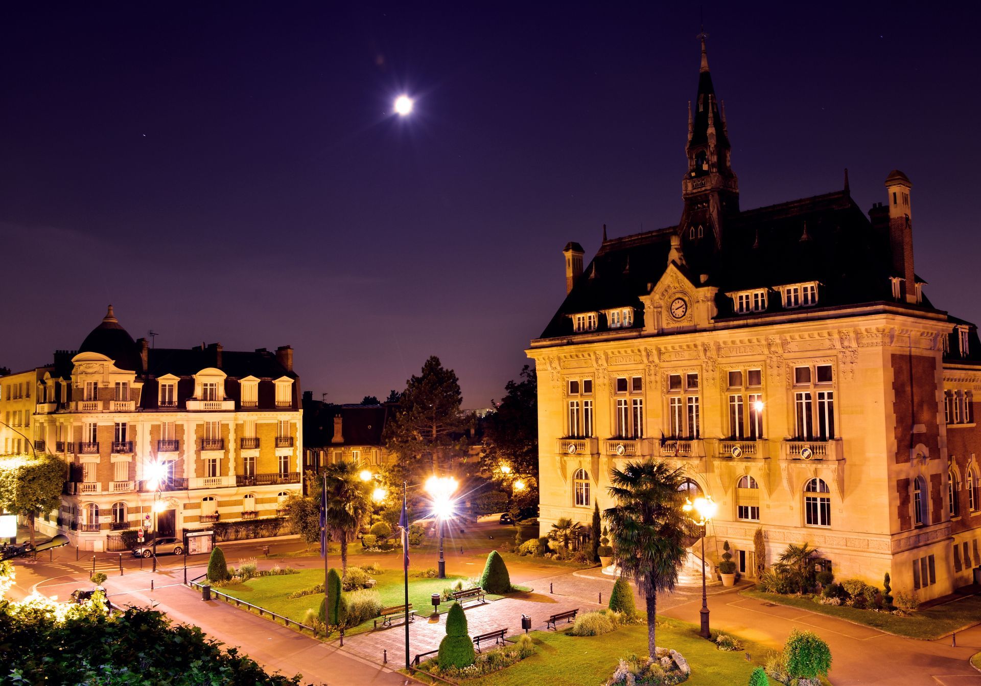 Vue nocturne de bâtiments illuminés sur une place de la ville sous un ciel éclairé par la lune.