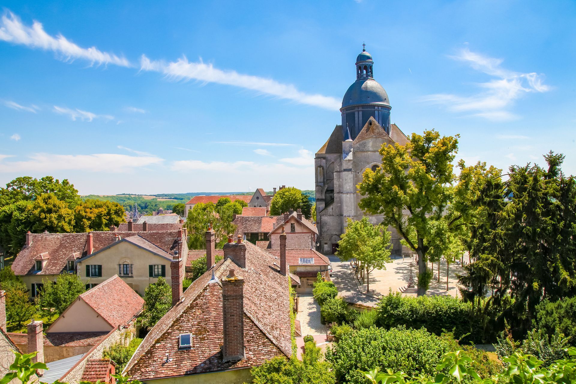 Vue d'une ville aux toits de tuiles rouges, d'une grande église à dôme et d'une végétation luxuriante sous un ciel bleu.