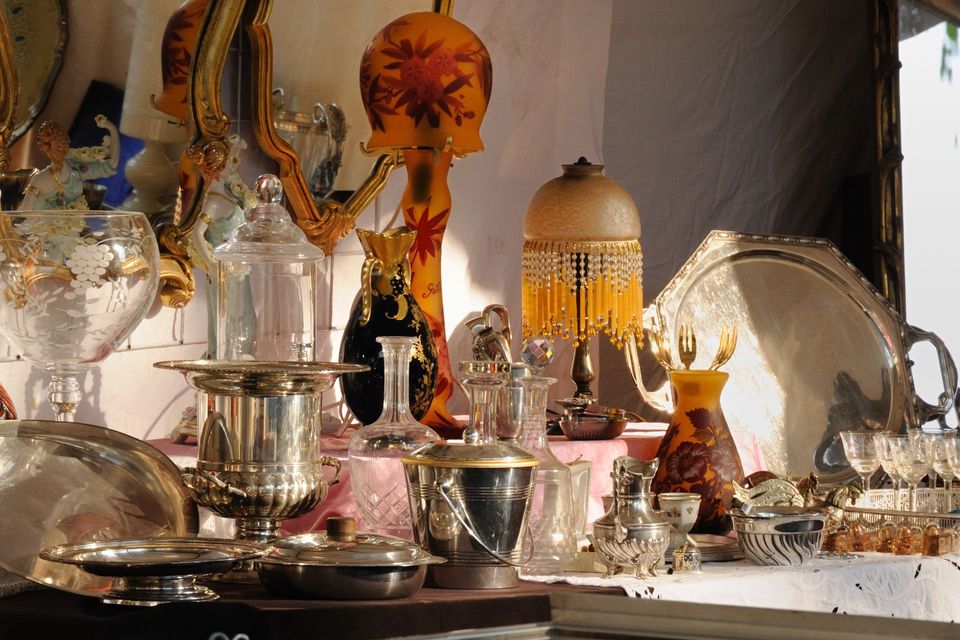 Présentation d'argenterie ancienne, de verrerie et de lampes sur une table devant une fenêtre.
