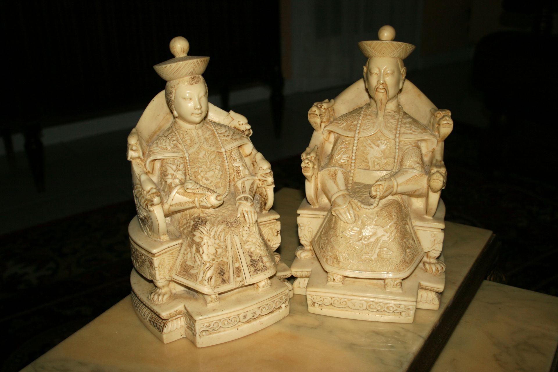 Deux statues sculptées en ivoire représentant des personnages assis, vêtus de robes et de chapeaux richement ornés.