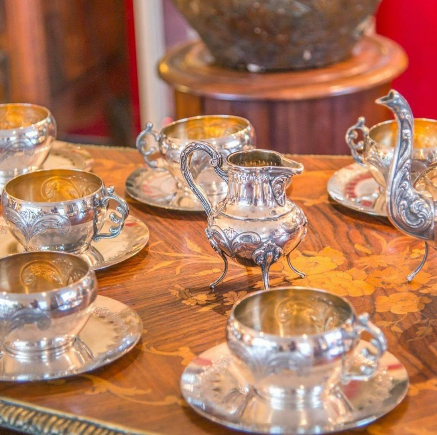 Tasses et soucoupes en argent sur une table en bois, avec un pot à lait et des éléments décoratifs.