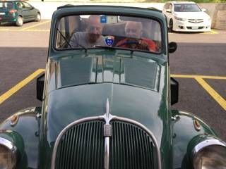 Voiture verte vintage avec deux personnes à bord, garée sur un parking délimité par des lignes jaunes.