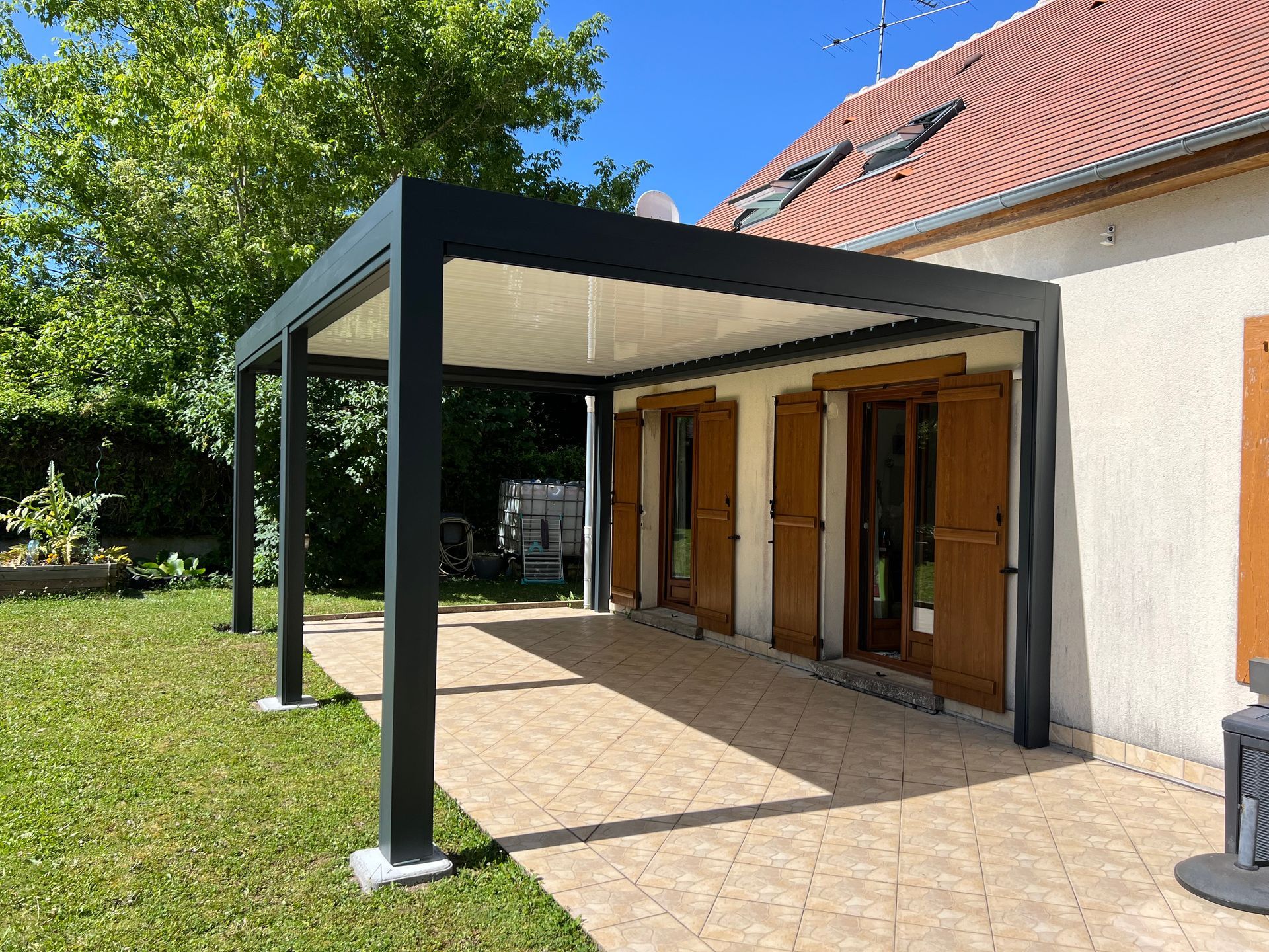 Une pergola noire est adossée à une maison aux volets bruns.
