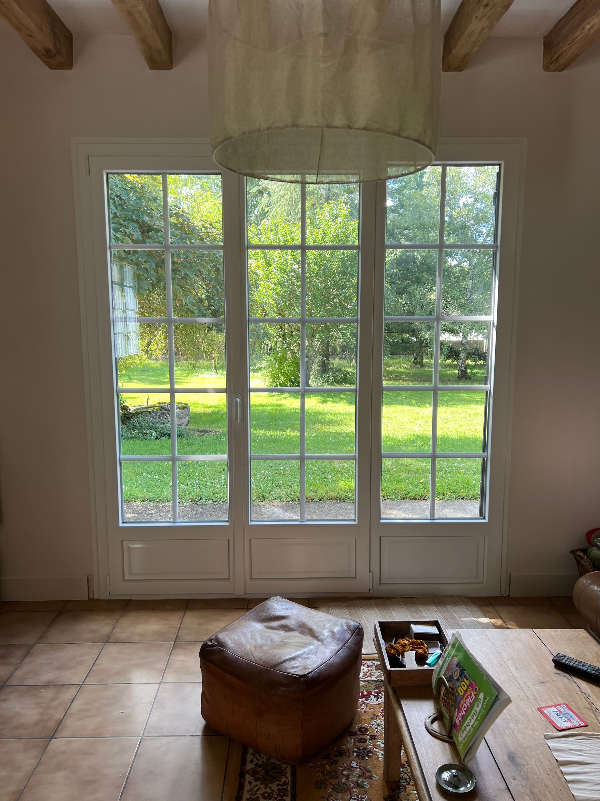 De grandes portes vitrées à cadre blanc donnent sur un jardin verdoyant, avec un pouf en cuir marron dans un salon carrelé.