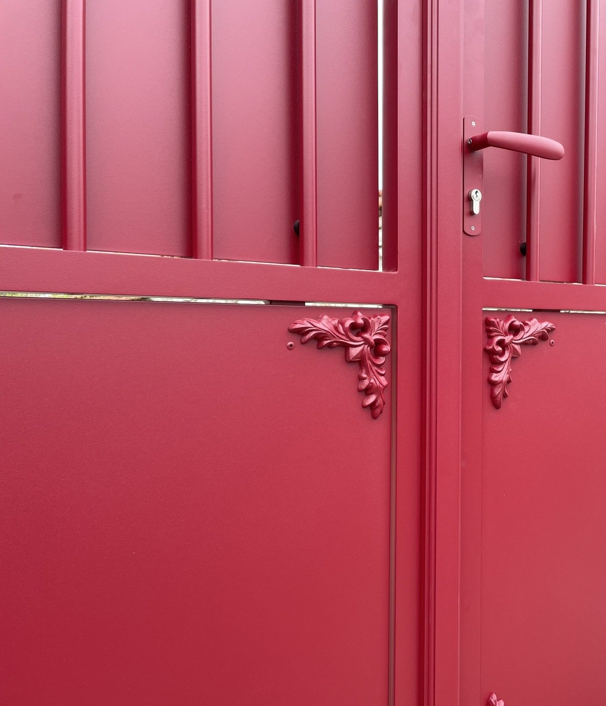 Portail en métal bordeaux orné de volutes décoratives, de filigranes d'angle et d'une simple poignée à levier.