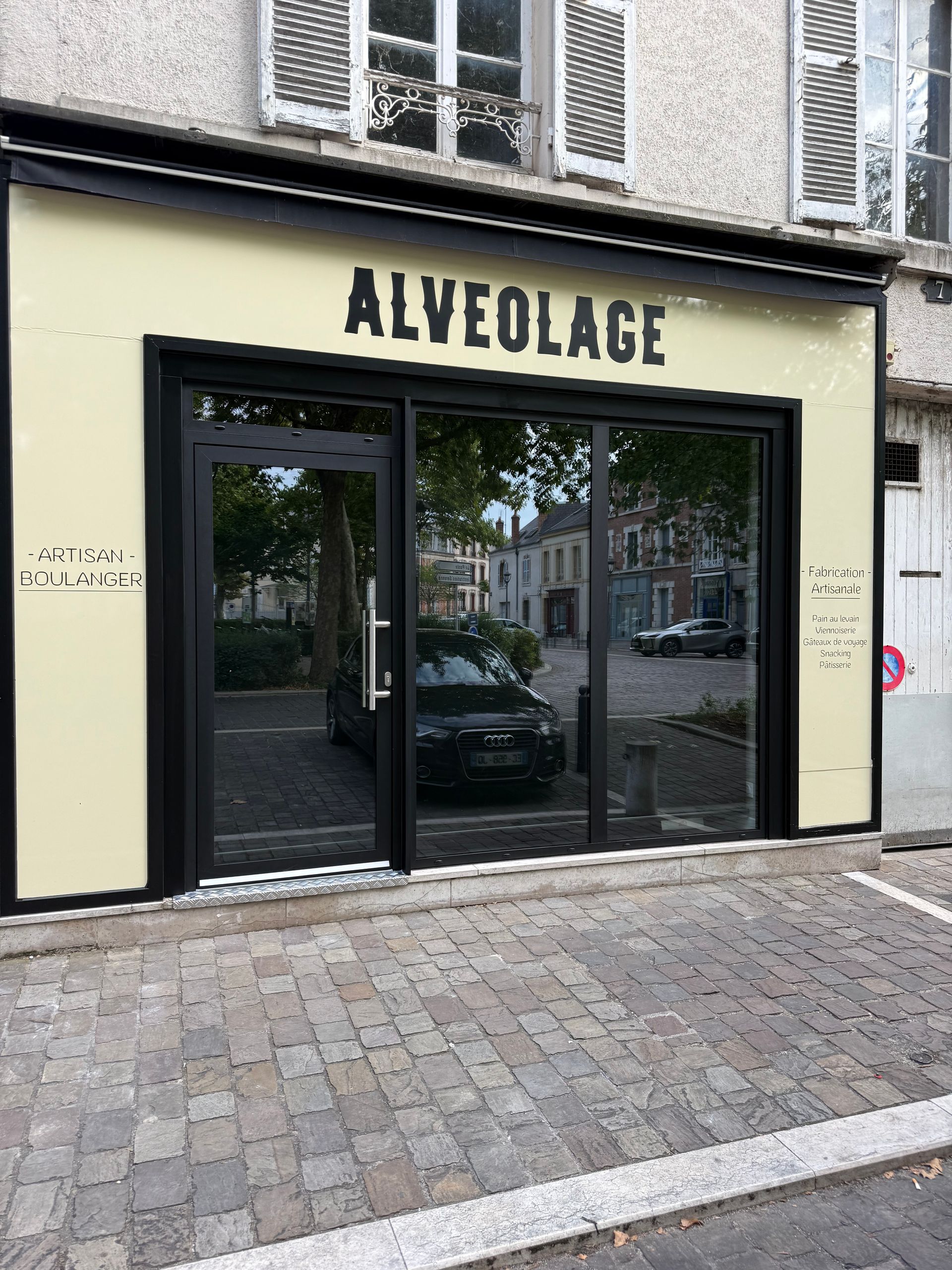 Une façade couleur crème avec un cadre noir et le mot ALVEOLAGE en lettres noires grasses au-dessus d'une grande vitrine.