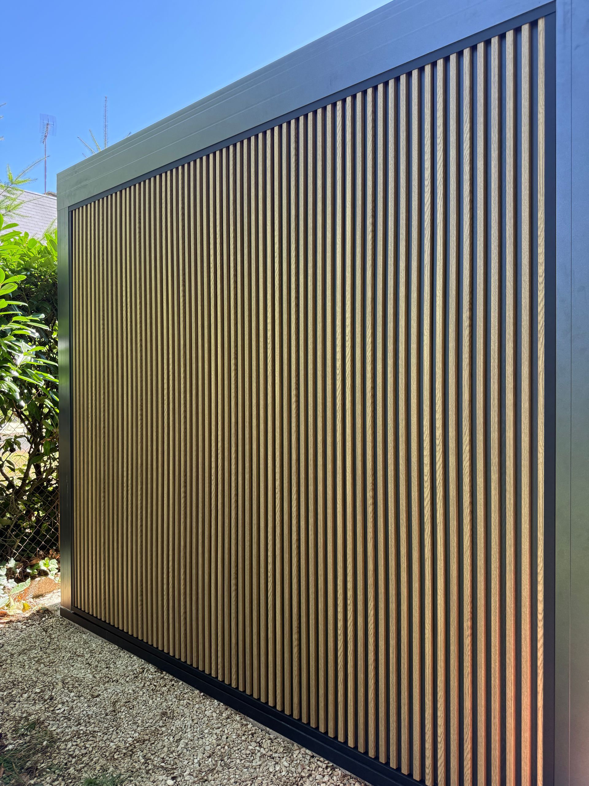 Un mur à cadre noir orné de nombreuses lattes de bois verticales de couleur claire, dans un jardin extérieur.
