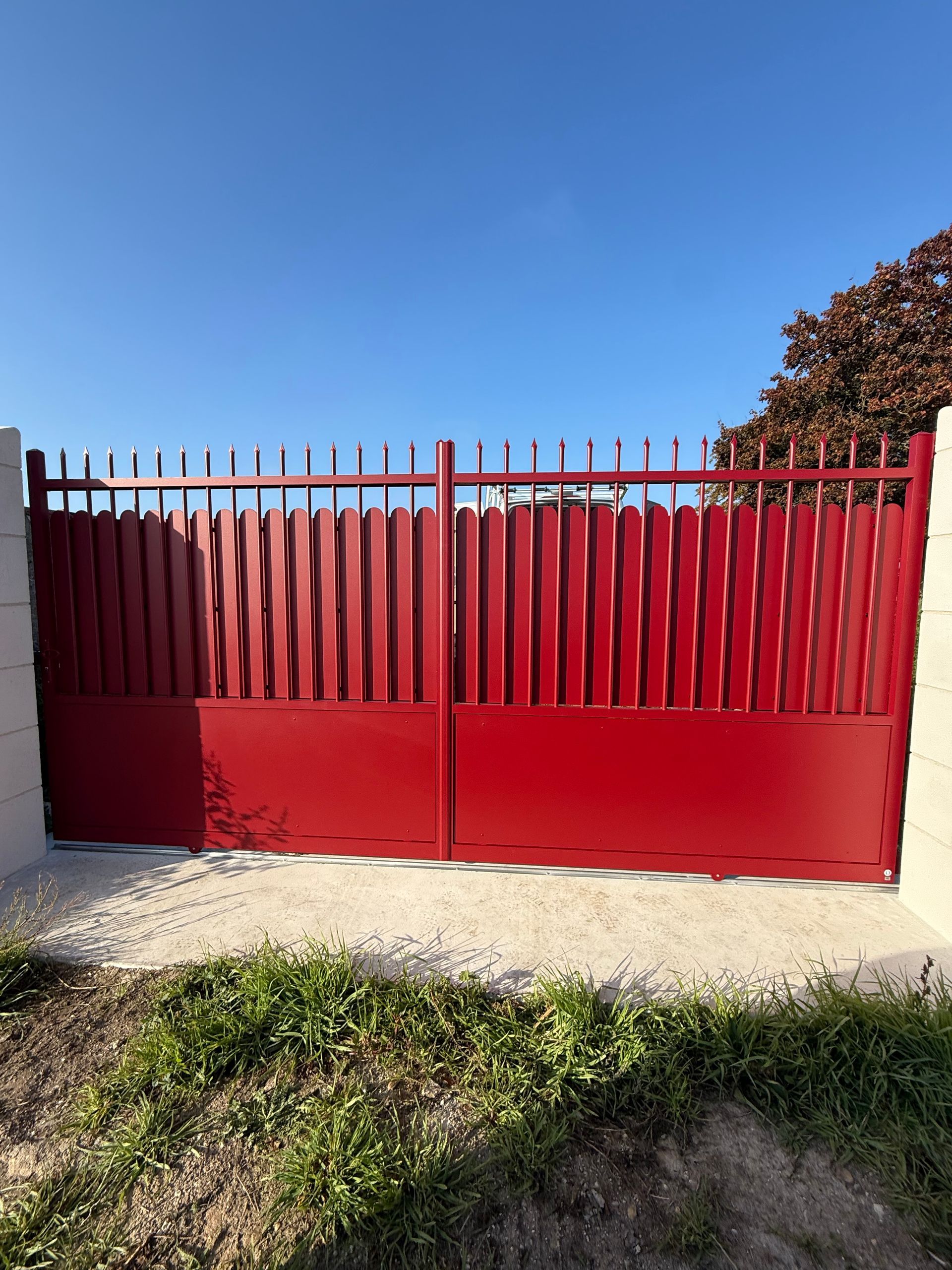Portail métallique rouge à barreaux verticaux, scellé dans du béton, sur fond de ciel bleu.
