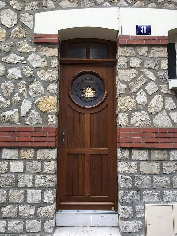 Une porte en bois brun avec une fenêtre circulaire est encastrée dans un mur de pierre, surmontée d'un panneau bleu numéro 8.