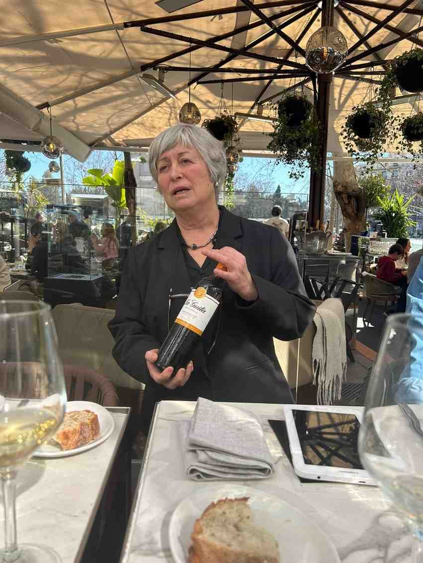 Una persona con una chaqueta negra sostiene una botella de vino en una mesa puesta para una comida.