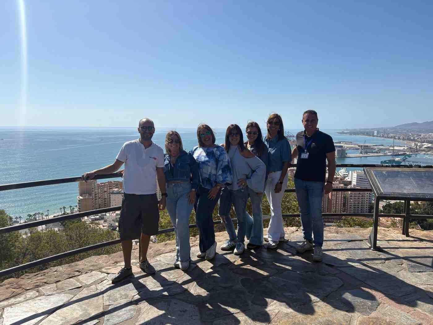 Un grupo de siete personas se encuentra en fila al aire libre, sonriendo a la cámara con vistas a una ciudad costera.