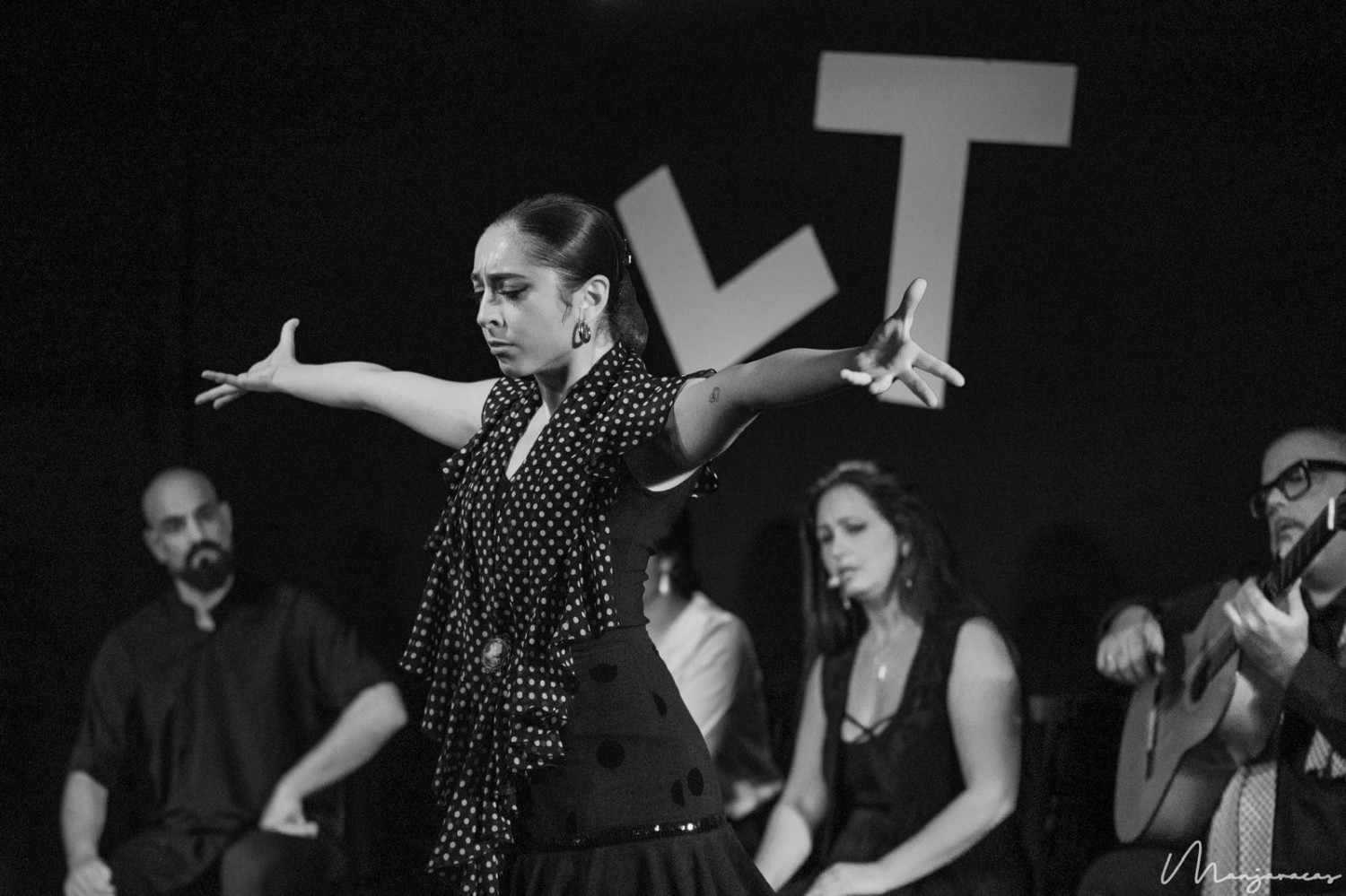 Una artista baila flamenco en el escenario con los brazos extendidos, acompañada por músicos sentados.