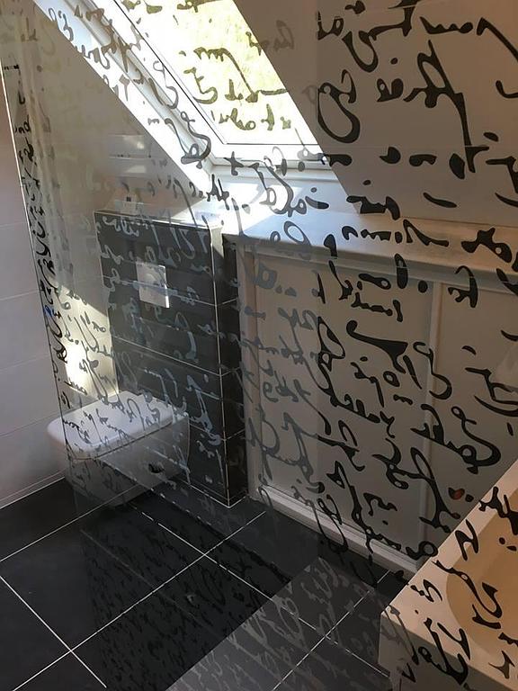 Petite salle de bains carrelée avec un espace douche, des carreaux de sol foncés et une cloison en verre dépoli avec des inscriptions arabes.