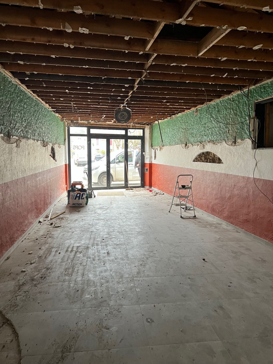 Longue pièce vide en cours de rénovation, avec plafond en bois apparent, deux chaises et des murs verts et roses près des portes vitrées.