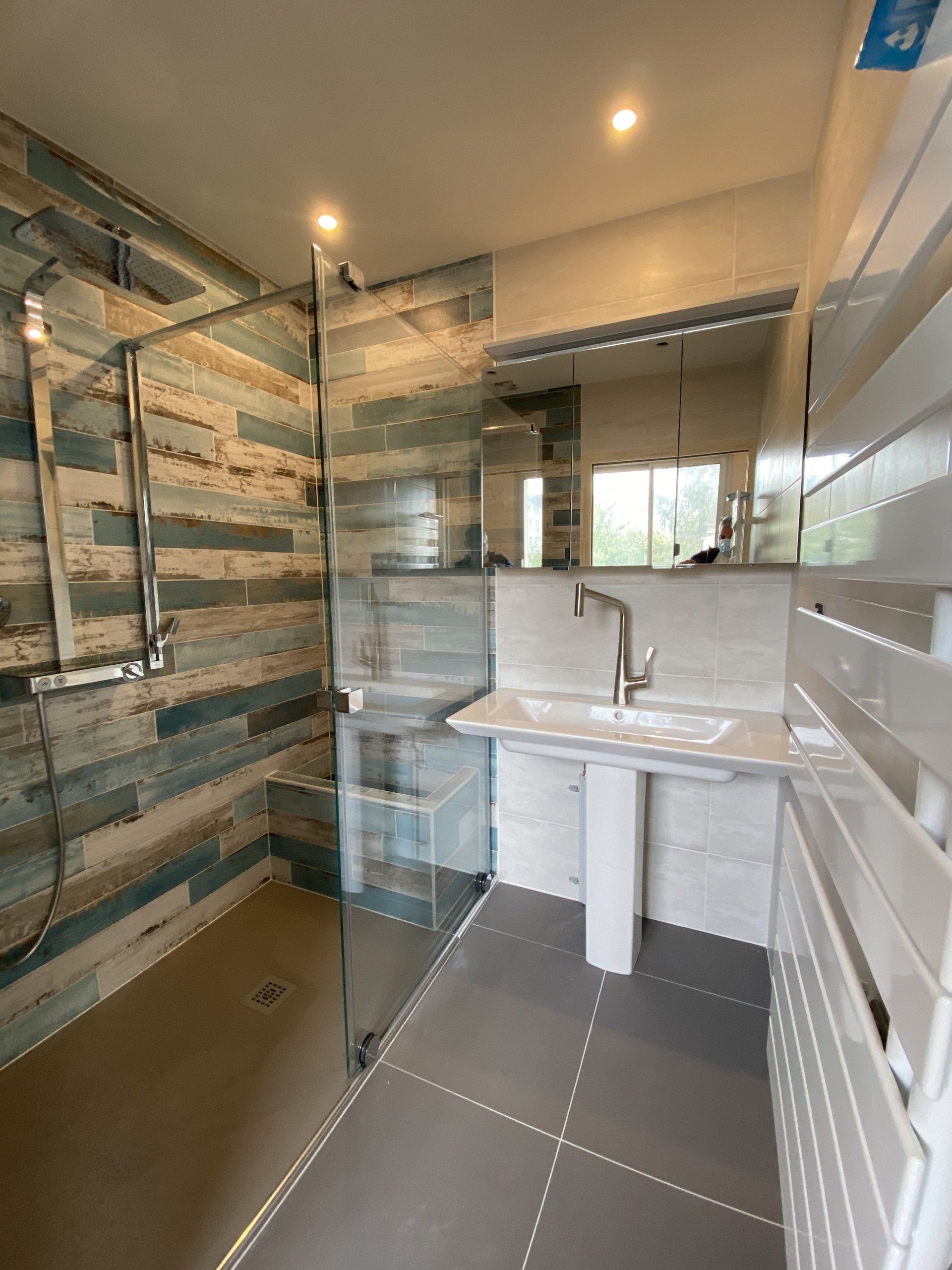Salle de bains moderne avec douche en verre, murs carrelés à rayures, meuble-lavabo blanc et grand miroir.