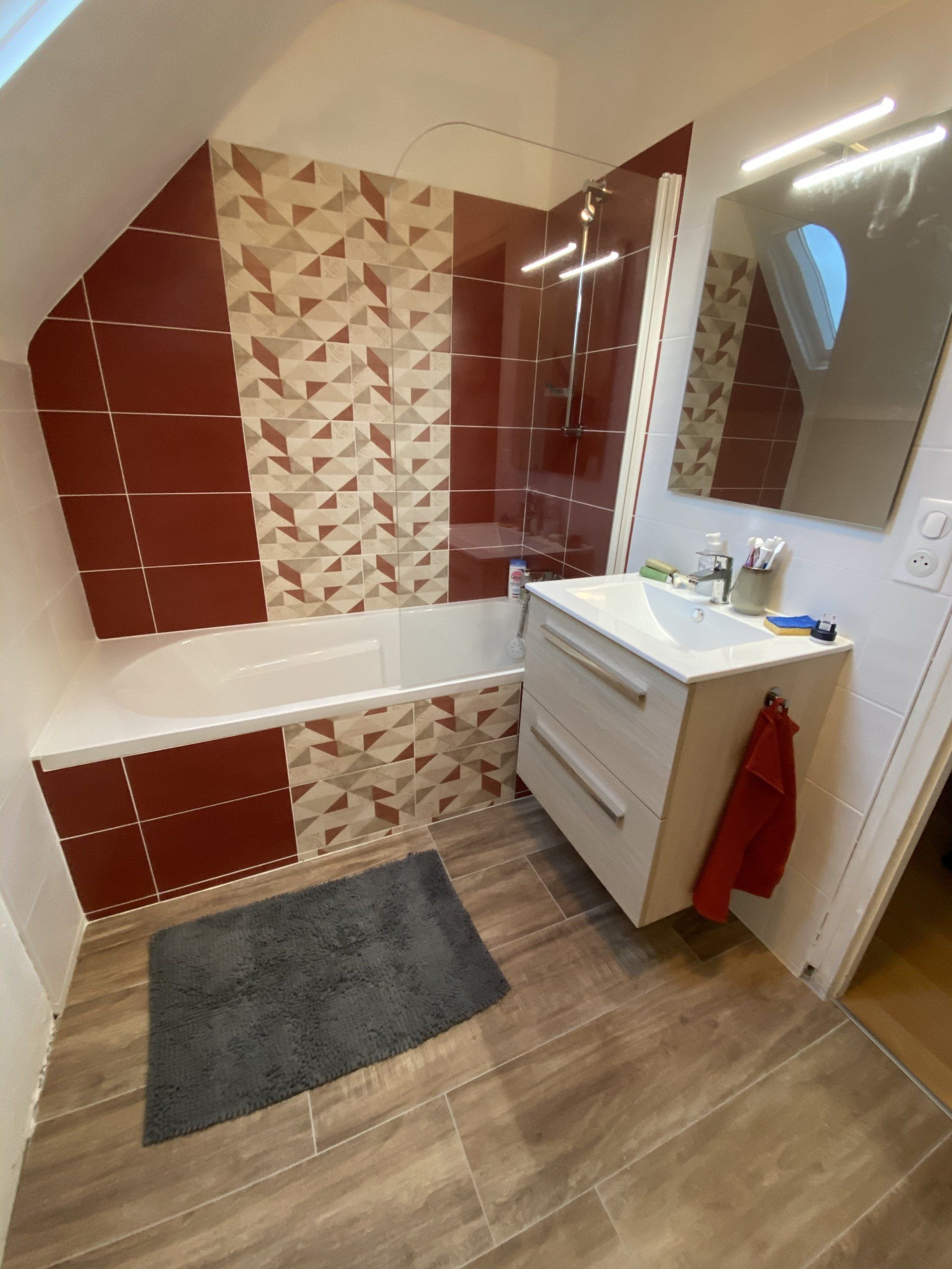 Petite salle de bains avec baignoire carrelée de rouge et beige, lavabo, miroir et tapis de bain gris.