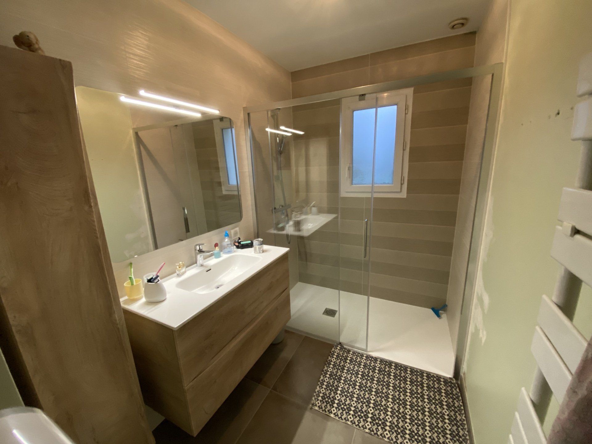 Salle de bains moderne beige avec meuble-lavabo suspendu en bois, douche à l'italienne, fenêtre et tapis de sol à motifs