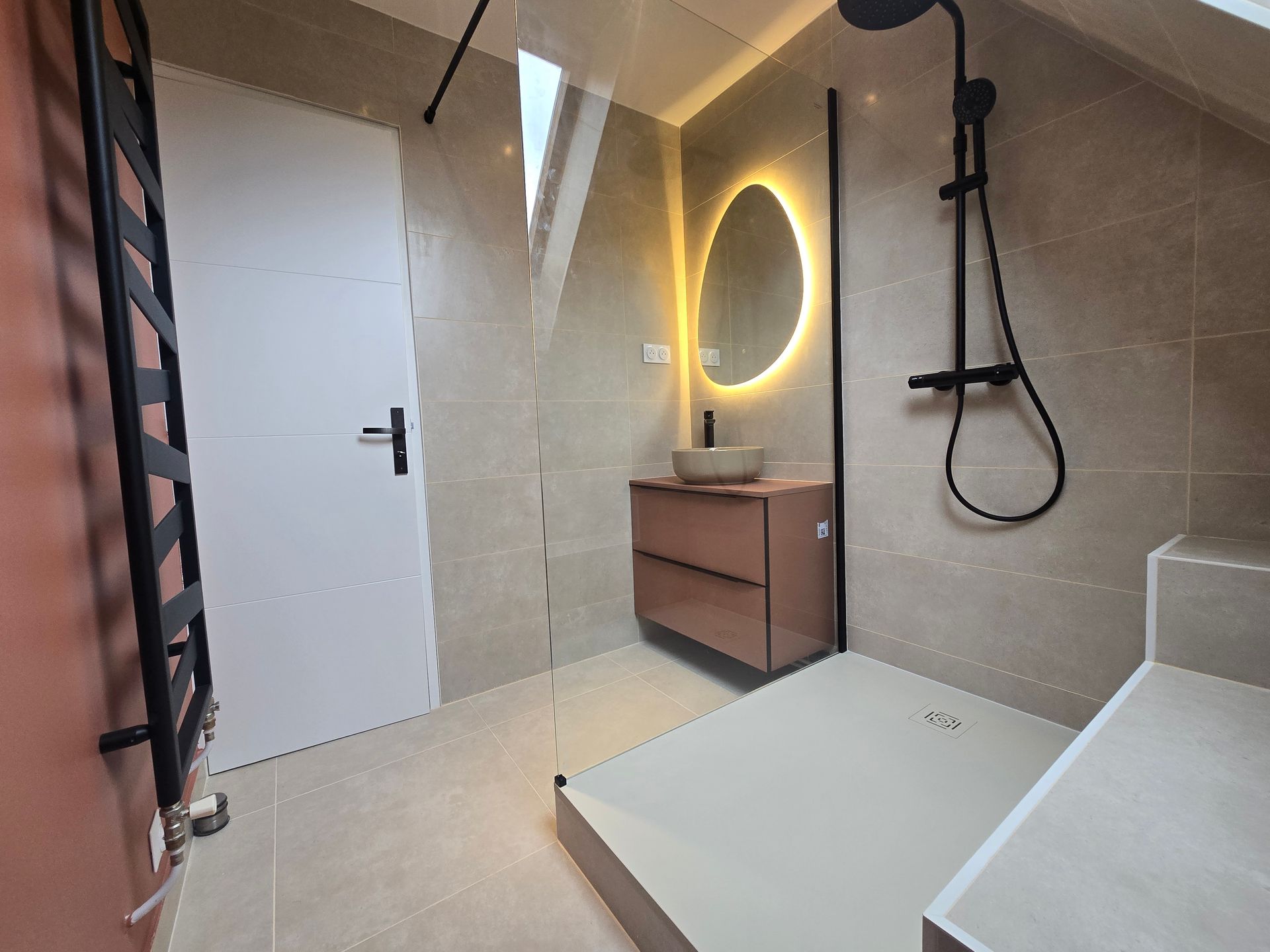 Une salle de bains moderne avec une douche à l'italienne, un lavabo suspendu avec miroir éclairé et un sèche-serviettes noir.