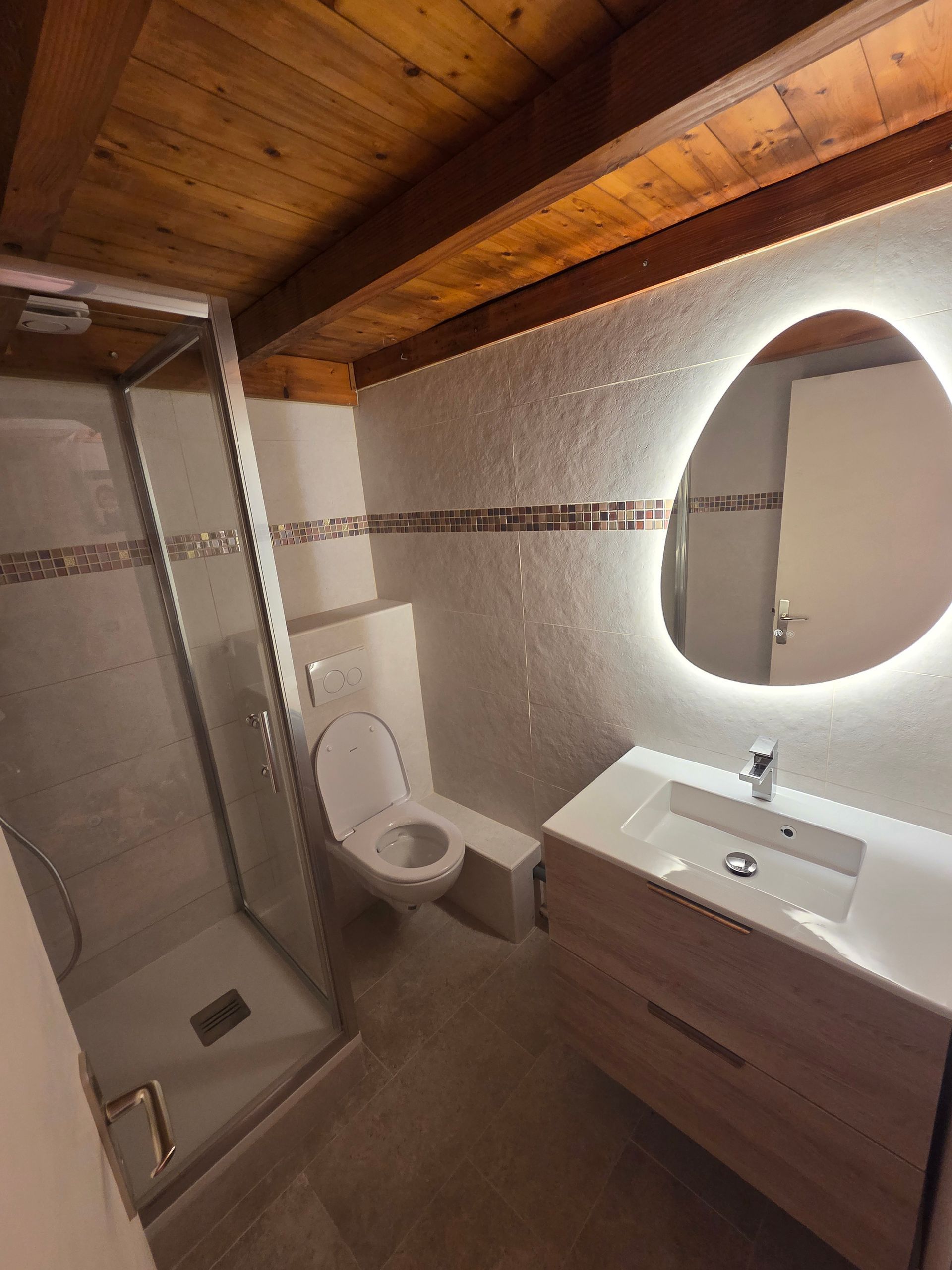 Salle de bains moderne avec douche vitrée, toilettes suspendues, lavabo et miroir éclairé aux formes organiques.