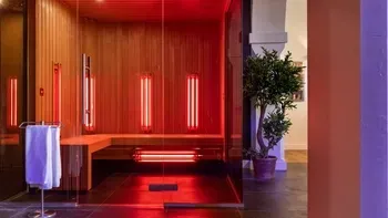 Rood verlichte infraroodsauna met houten wanden, banken, rode verwarmingselementen, een handdoekenrek en een potplant buiten.