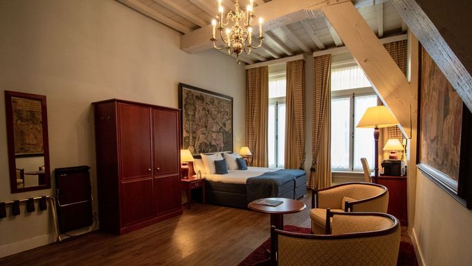 Hotelkamer met bed, houten meubilair, ramen en kroonluchter.