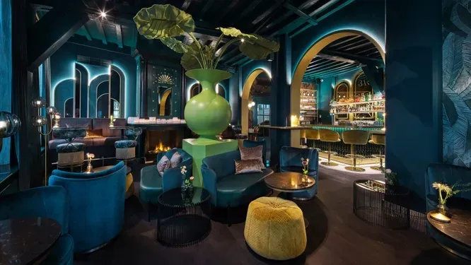 Luxe blauwgroene bar met zachte stoelen, een grote groene vaas en gewelfde deuropeningen.