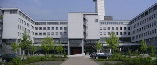 Großes, weißes, mehrstöckiges Gebäude mit zentralem Eingang, Gehweg und Bäumen davor
