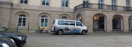 Ein weißer Lieferwagen parkt vor einem beigen Gebäude mit gewölbten Türen und Fenstern; daneben steht ein Fahrrad.