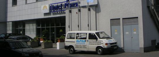 Ein weißer Lieferwagen mit blauer Beschilderung parkt vor dem Eingang eines Gebäudes, daneben steht ein dunkles Auto.