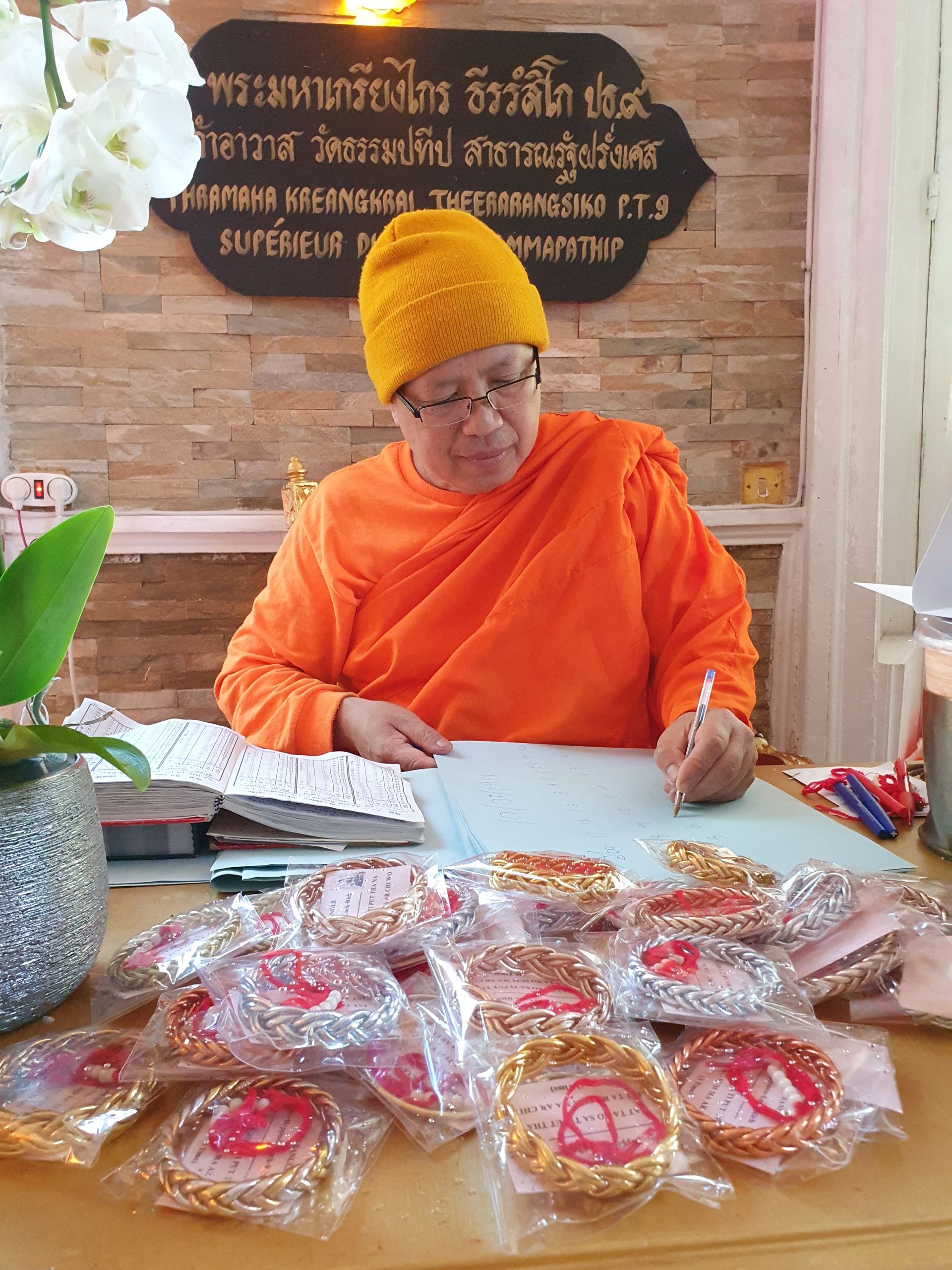 Un moine en robe orange est assis à un bureau, entouré de bracelets emballés, en train d'écrire.