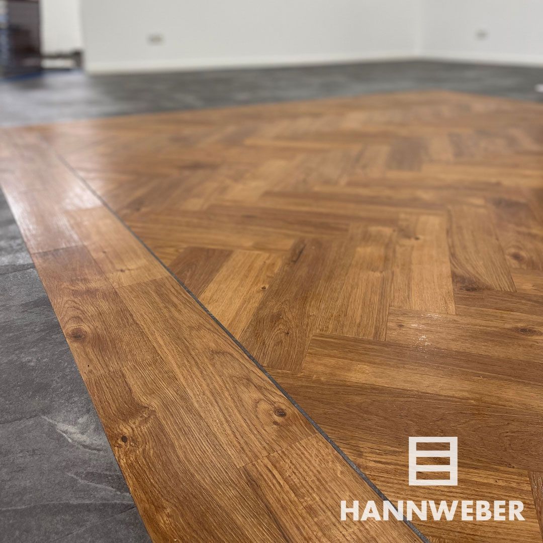 Nahaufnahme eines Fischgrätparketts aus Holz, eingefasst von dunkelgrauen Fliesen, in einem leeren Raum
