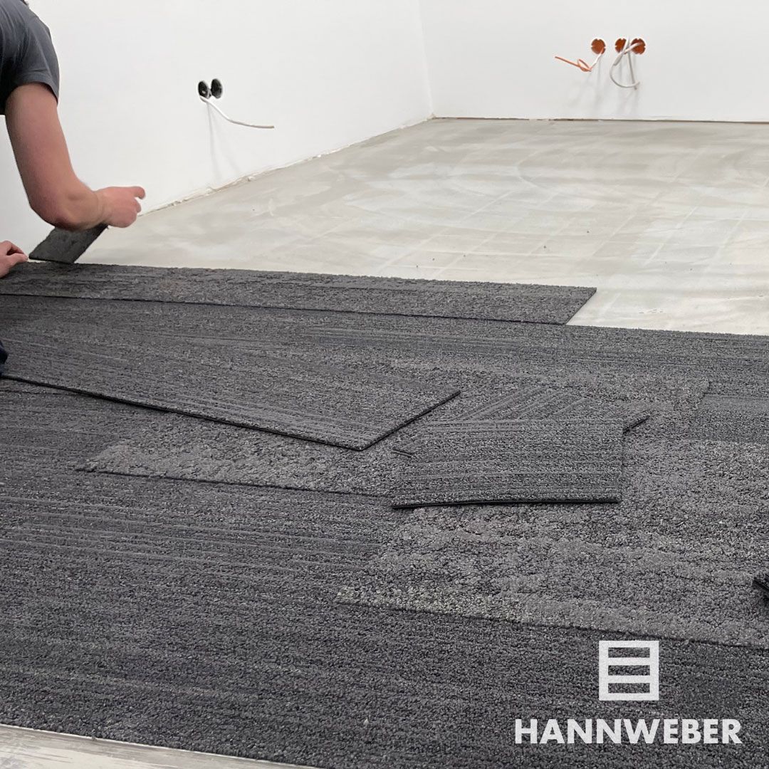 Eine Person verlegt dunkelgraue Teppichfliesen in einem weißen Raum; im Vordergrund liegen lose Fliesen.