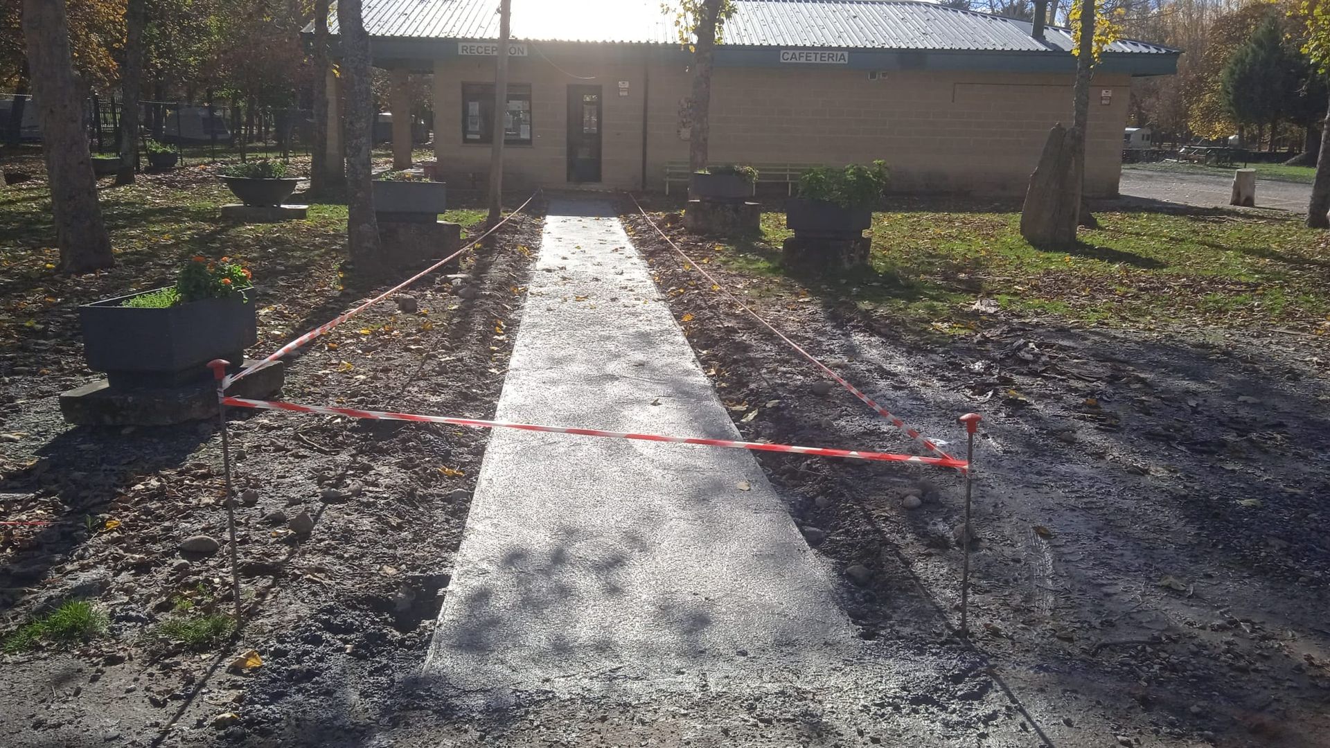 Camino de hormigón hacia el edificio, bloqueado con cinta roja y blanca. Hojas otoñales esparcidas.