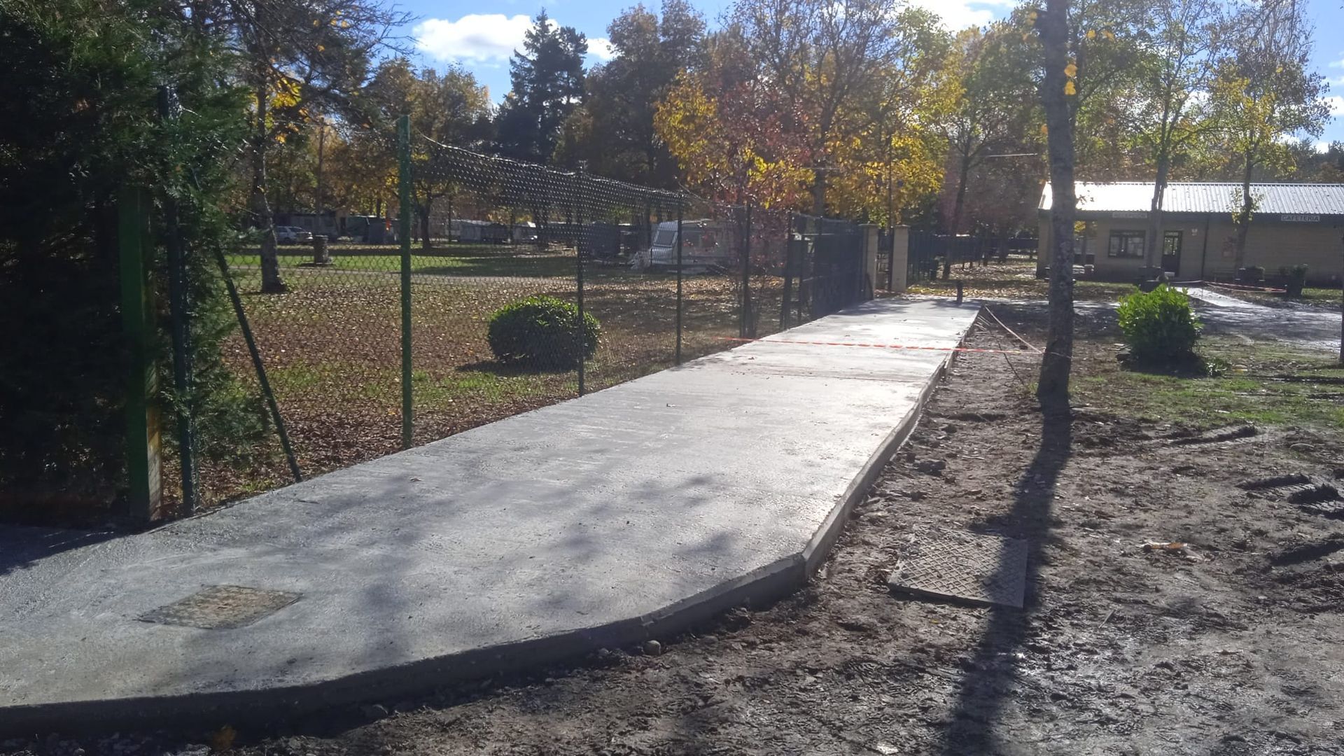 Camino de hormigón a través de un parque con una valla a la izquierda y árboles al fondo.