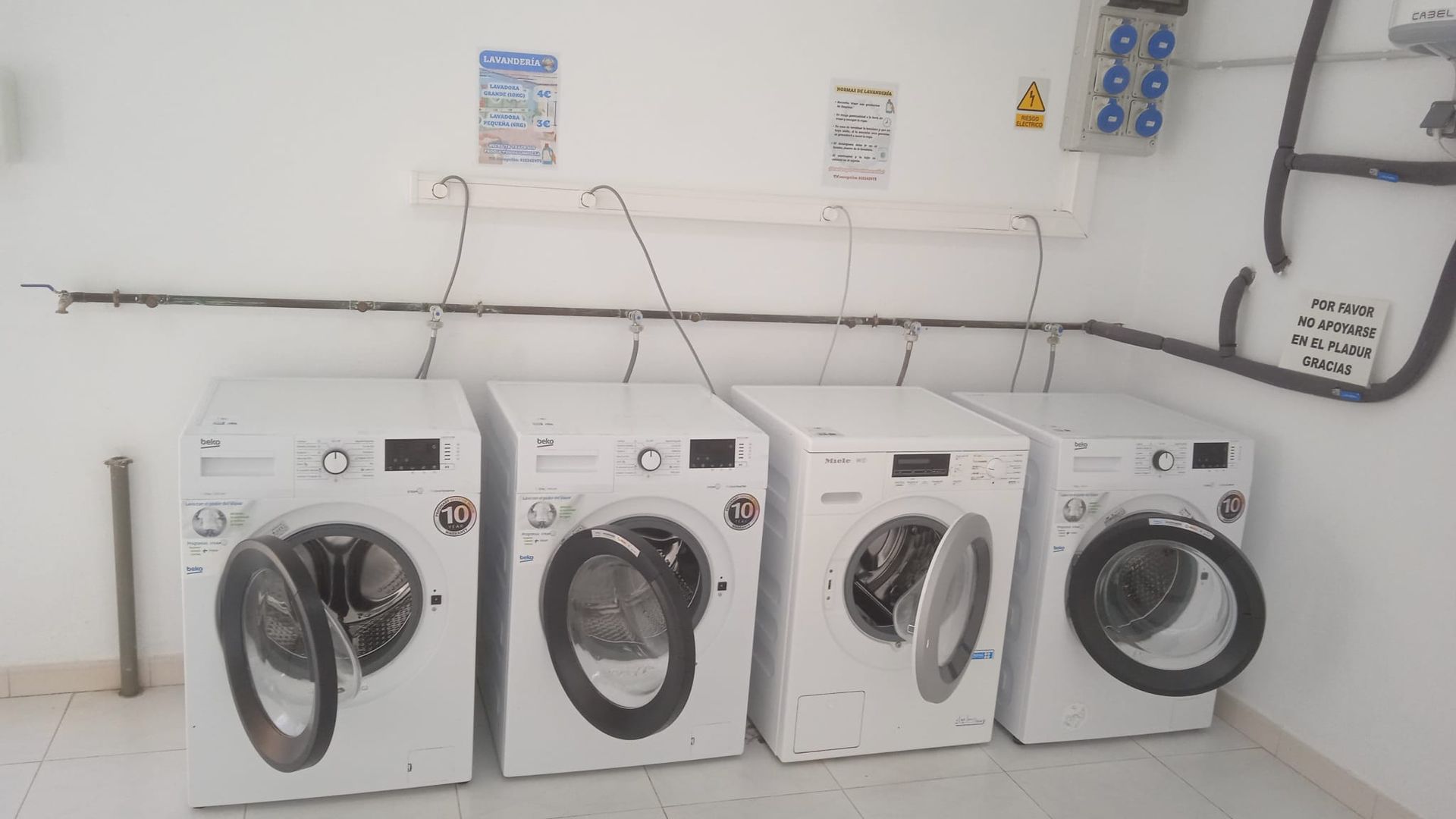 Cuatro lavadoras blancas con las puertas abiertas en un lavadero, conectadas a tomas de corriente.