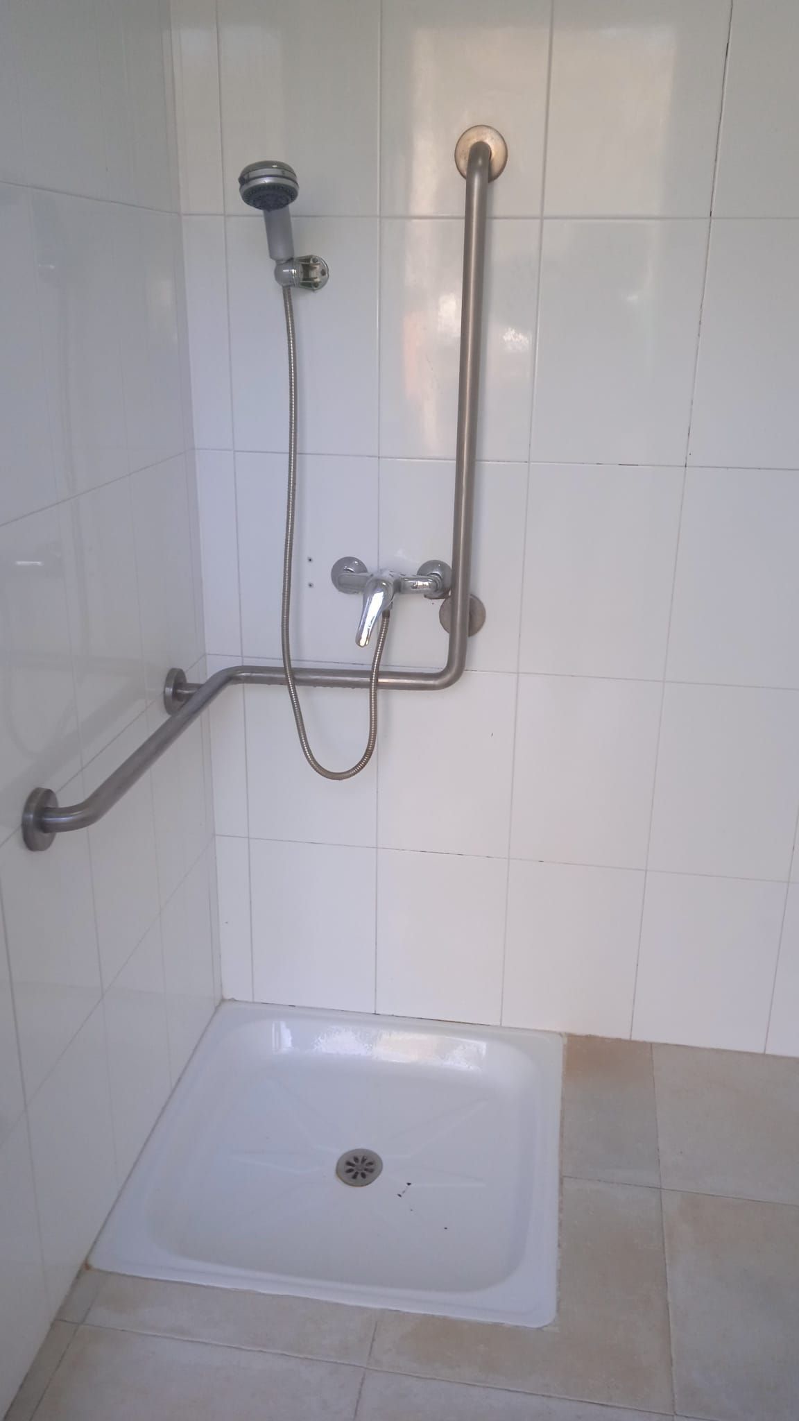 Ducha accesible con pasamanos, azulejos blancos y lavabo de acceso bajo.