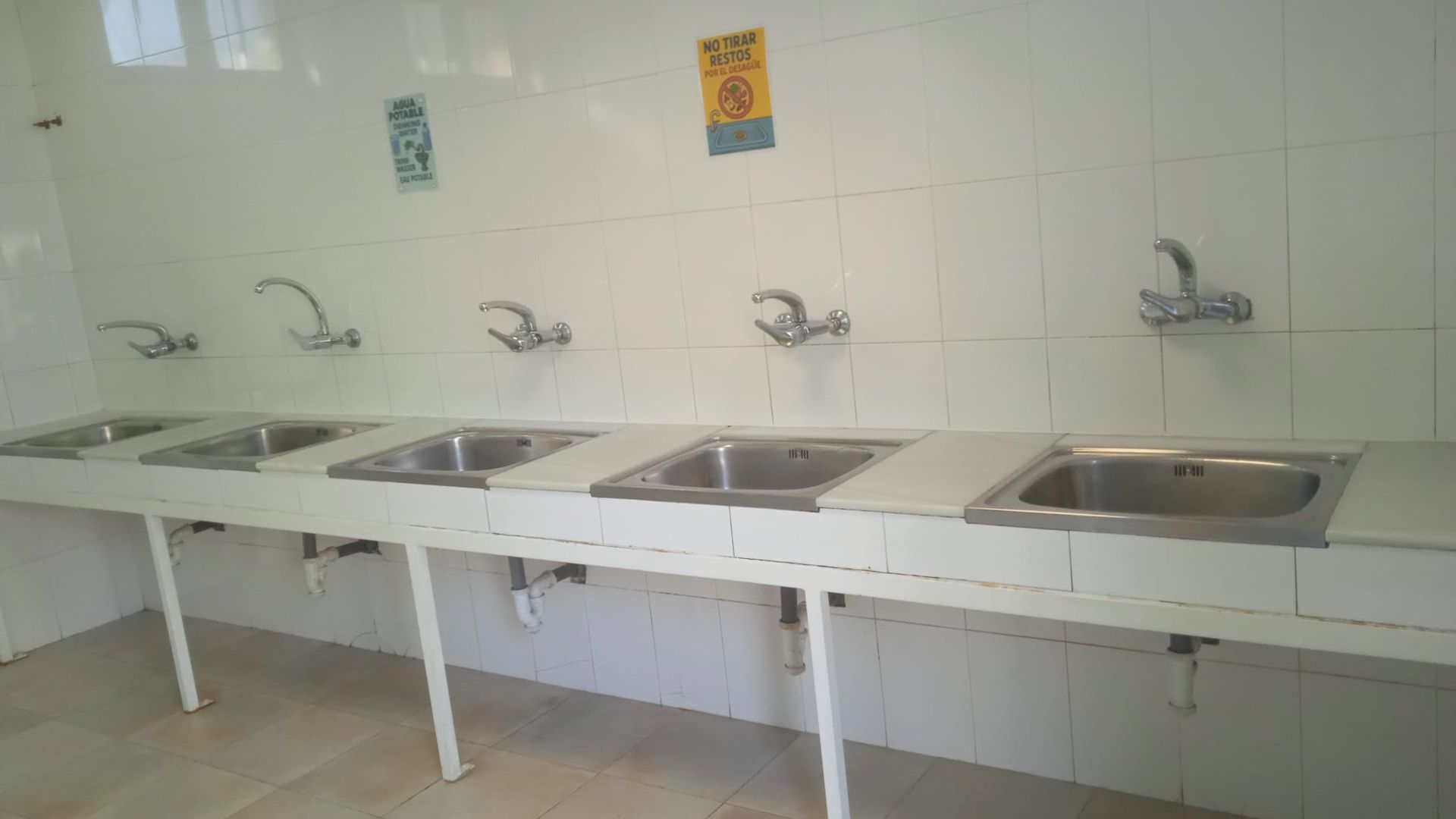 Fila de lavabos contra una pared de azulejos blancos con grifos de agua en un baño público.