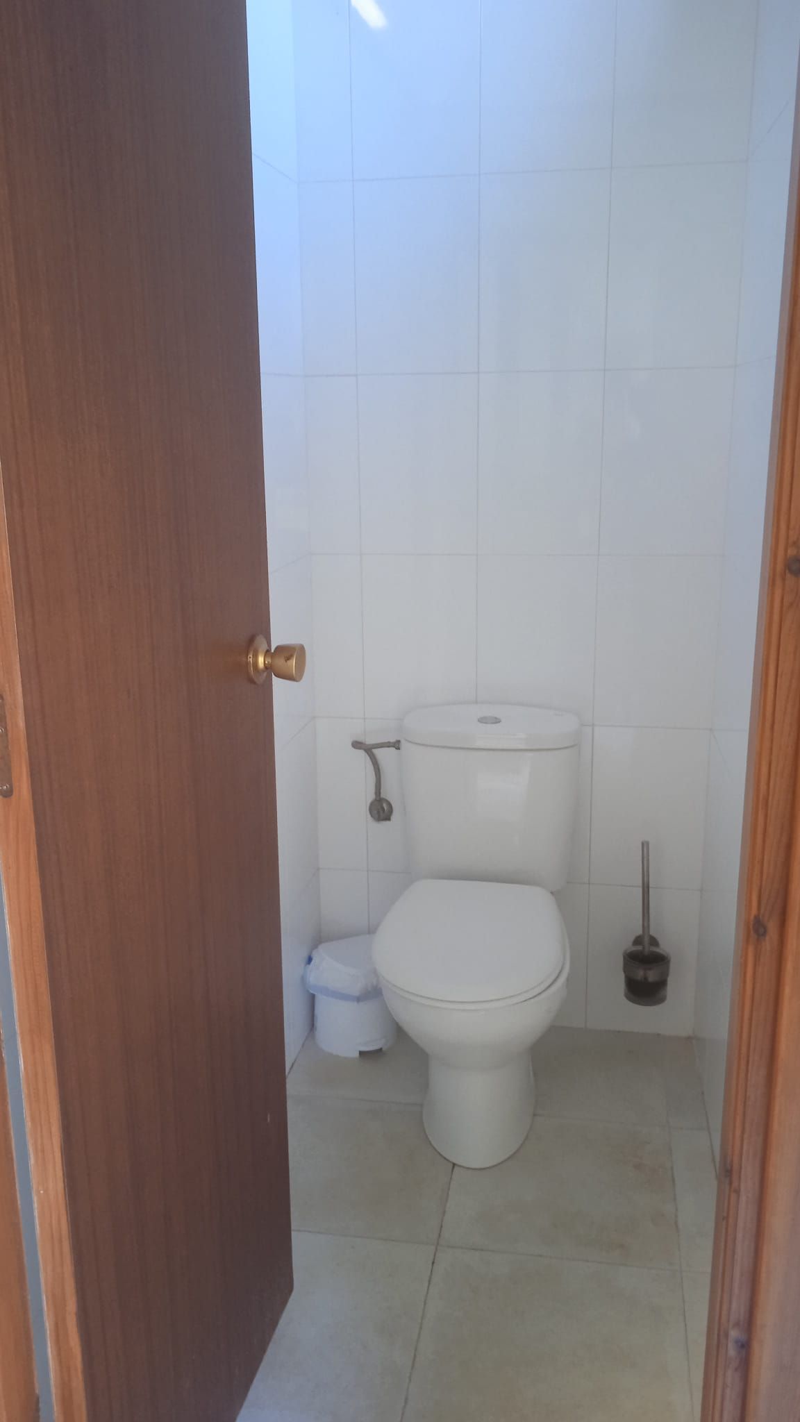 Pequeño baño con azulejos blancos, inodoro cerrado, papelera y escobilla. La puerta de la izquierda está entreabierta.