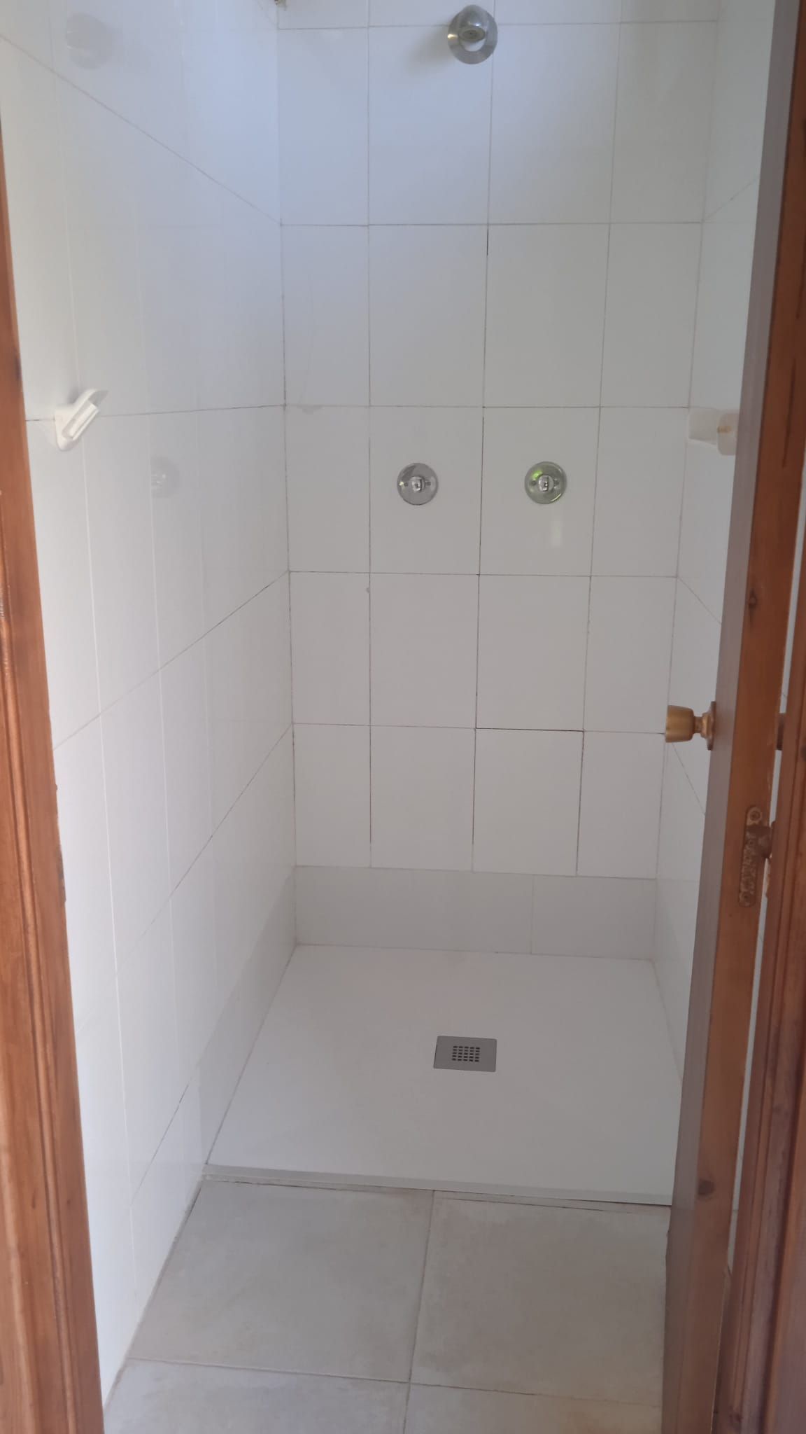 Ducha con azulejos blancos, dos grifos y desagüe. Marco de puerta de madera a ambos lados.