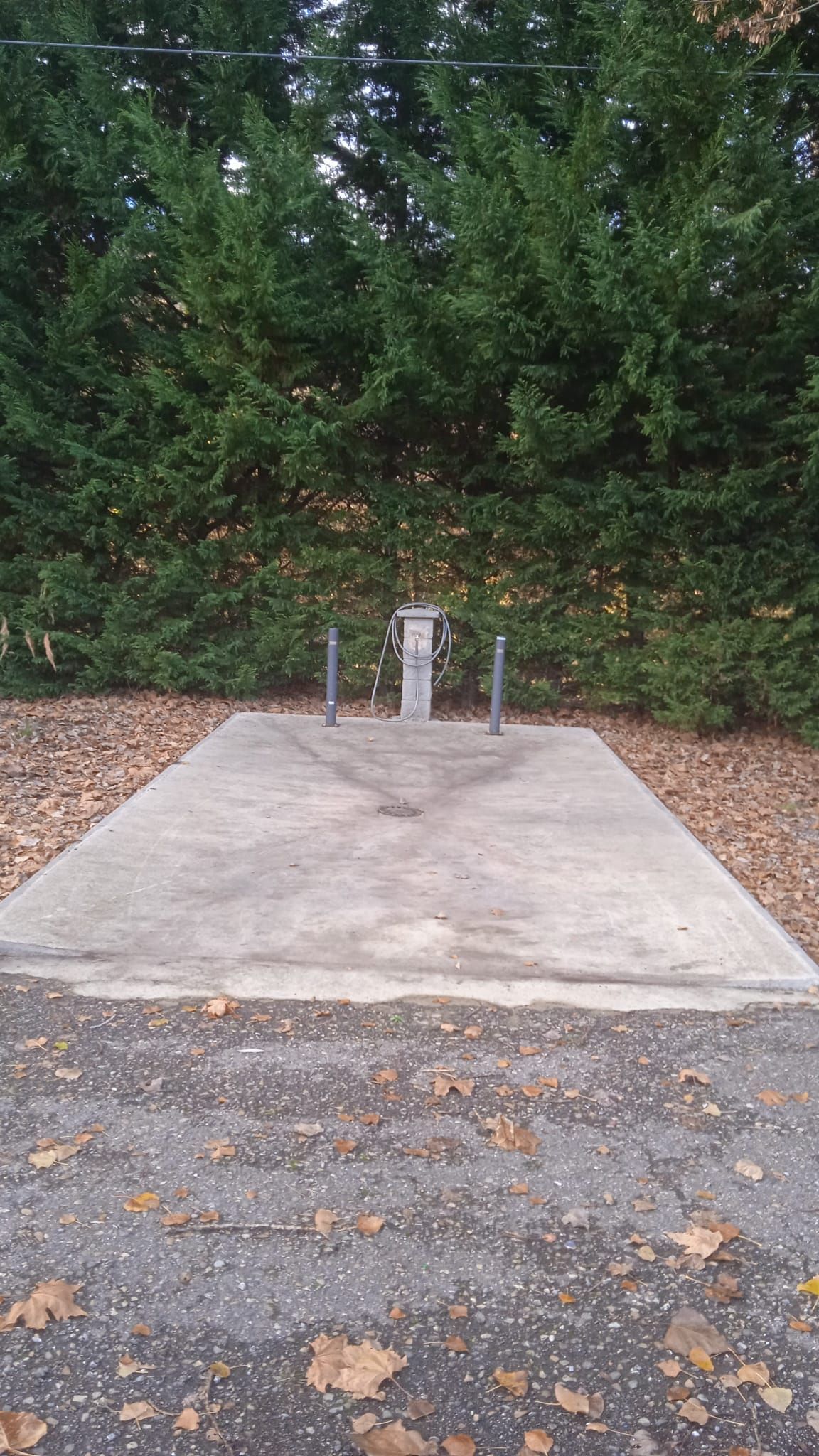 Losa de hormigón con grifo de agua y tuberías, delante de un seto y hojas esparcidas.