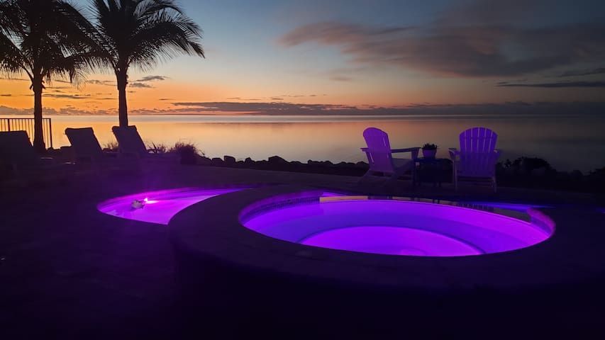 Two glowing purple hot tubs near a beach at sunset with two chairs.