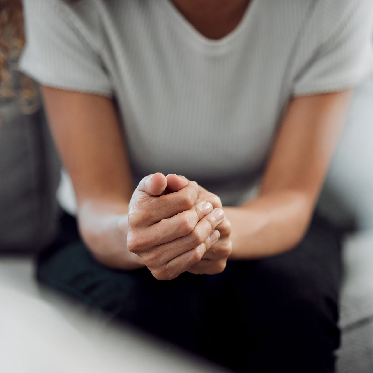 Personne assise, les mains jointes, portant une chemise gris clair et un pantalon noir.