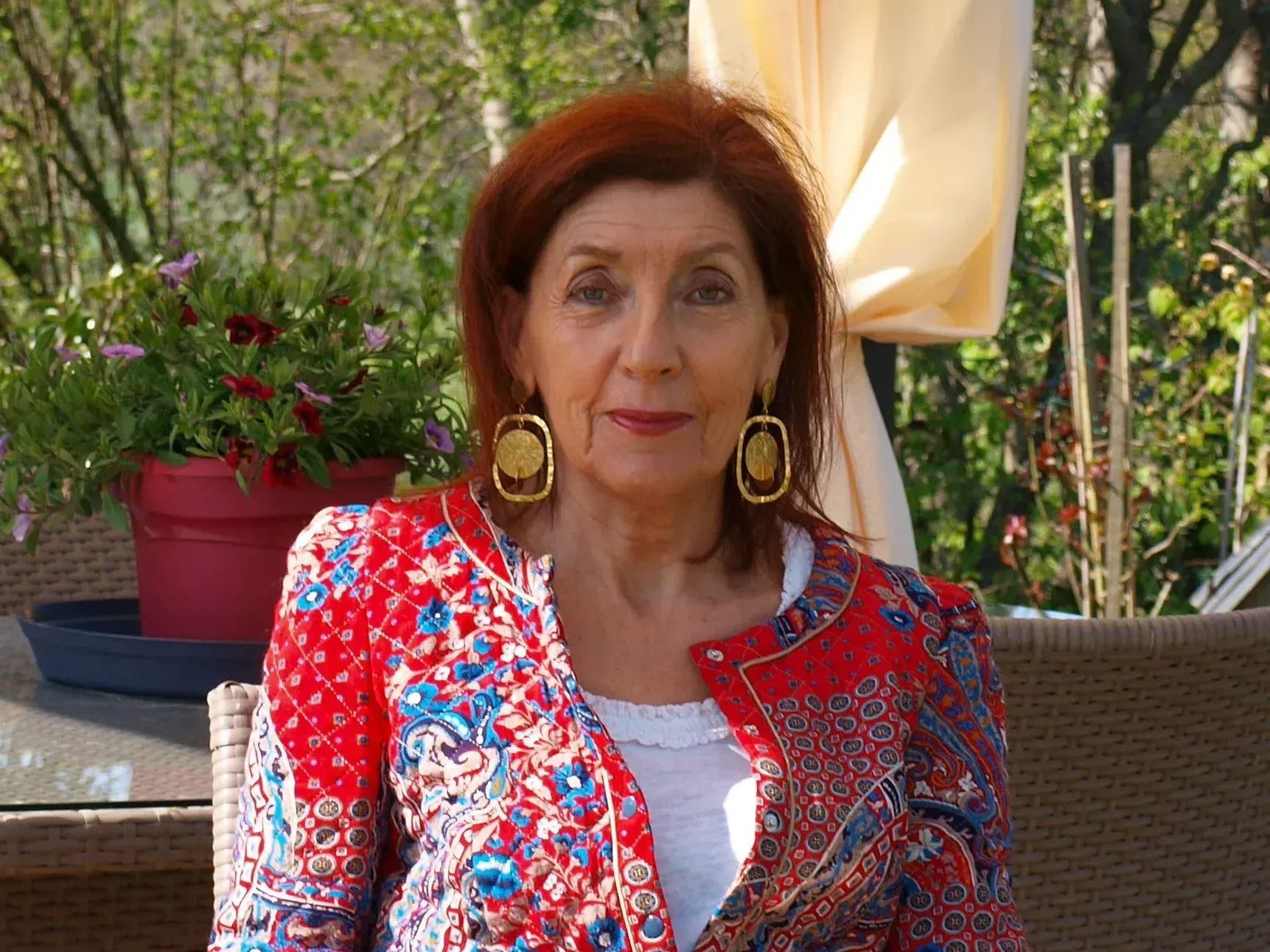 Femme assise en terrasse, vêtue d'une veste à motifs rouges et de boucles d'oreilles dorées.