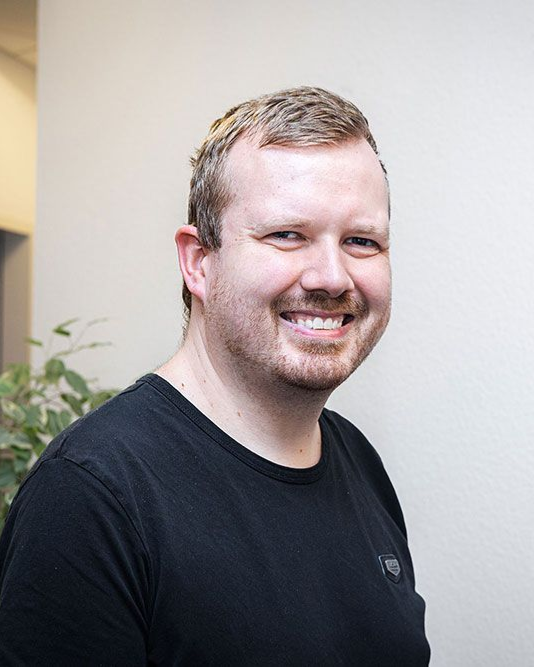 Ein Mann mit kurzen blonden Haaren und einem schwarzen T-Shirt lächelt in die Kamera; er steht vor einer weißen Wand.